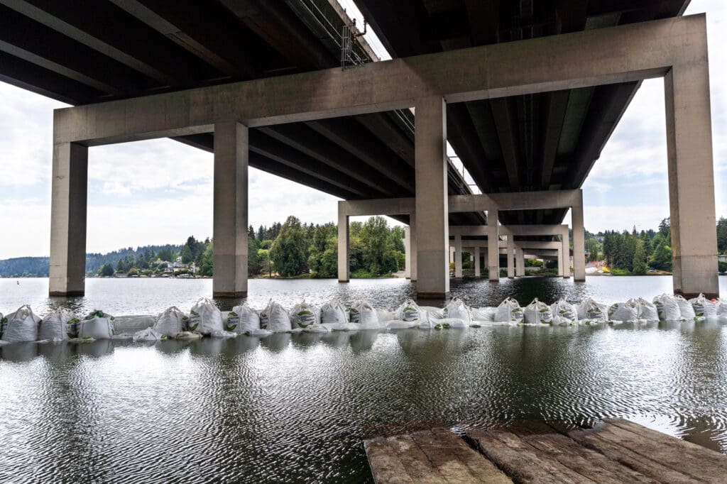 bags of sand are placed in a circle in the water next to the shoreline under the Bellevue side of the I-90 bridge. The concrete pillars of the bridge rise above the water, and the highway takes up most of the skyline.